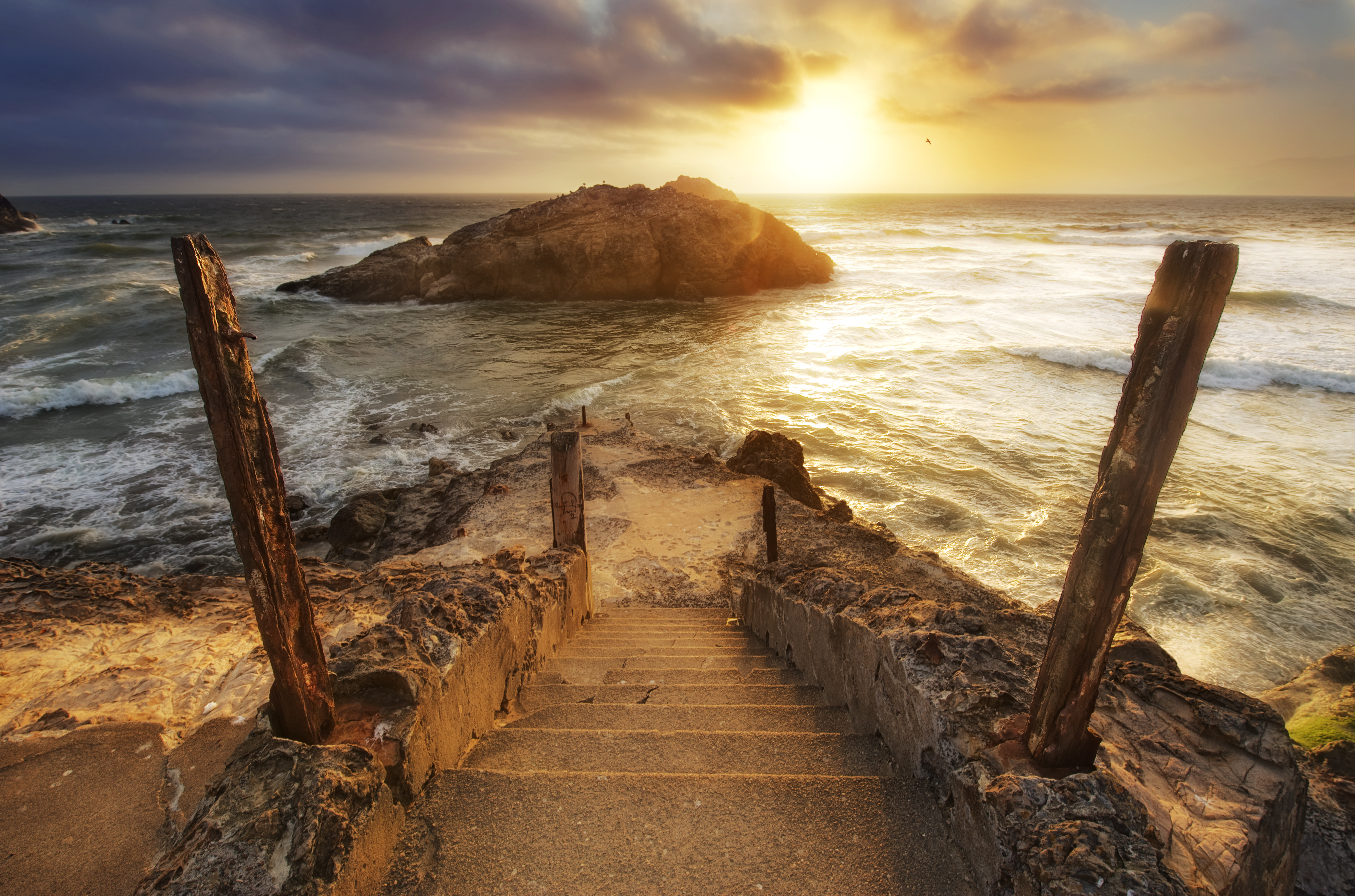 A beautiful sunset over calm seas, but with waves, rocks, and a rusty concrete staircase in the foreground.